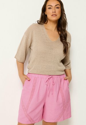 Kvinde iført en beige V-hals trøje med albuelange ærmer og lyserøde shorts med snøre og blonde detaljer, hænderne i lommerne, foran en hvid baggrund.