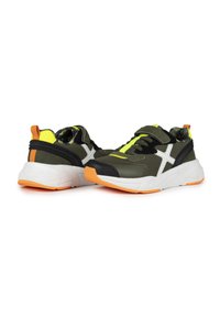 Zapatillas deportivas verdes con un diseño en forma de X blanca, detalles en negro, toques amarillos y suela naranja. Se observa una correa de velcro y un material texturizado.