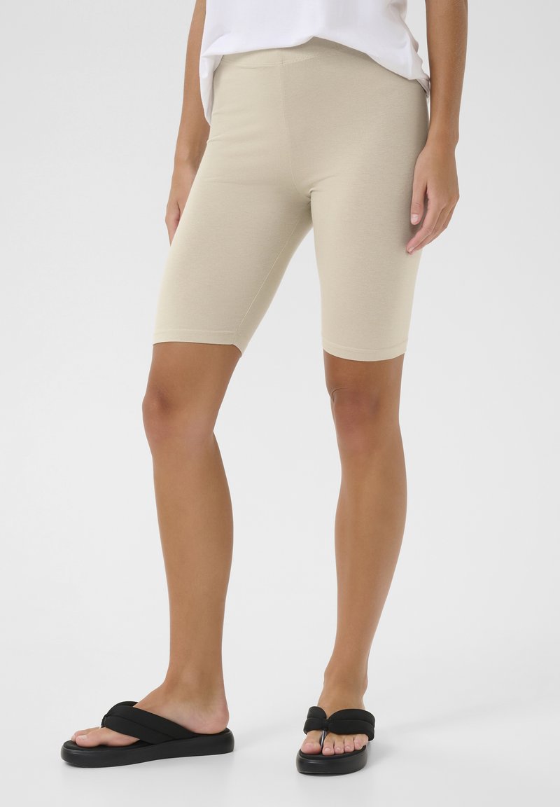 Beige cykelshorts lavet af blødt stof, med en tætsiddende pasform og midt på låret længde, parret med sorte flip-flops.