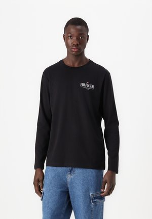 Schwarzes Langarmshirt aus Baumwolle mit rundem Halsausschnitt und Front-Logo. Kombiniert mit hellblauen Jeans.