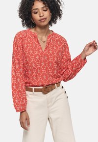 Blusa roja con estampado, de manga larga y escote en V, que presenta diseños florales blancos. Combinada con pantalones beige de talle alto y un cinturón marrón.