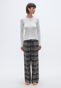 Femme debout pieds nus, portant un haut à manches longues gris clair et un pantalon de pyjama à carreaux gris avec de petits motifs subtils.