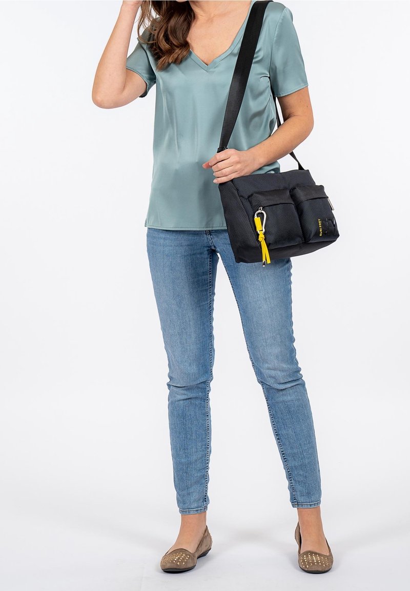 Czarna torebka o teksturowanej surface z żółtym uchwytem na suwak i podwójnymi przednimi kieszeniami, noszona na krzyż. Modelka ma na sobie turkusową bluzkę i jasne dżinsy.