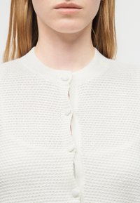 Γυναίκα που φοράει λευκό πλεκτό ζακέτα με υφή, κουμπωμένη στην κορυφή, δείχνοντας μέρος του λαιμού και της ωμοπλάτης.