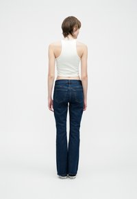 Osoba z krótkimi włosami ubrana w białą koszulkę bez rękawów, ciemnoniebieskie dżinsy i białe trampki, stoi tyłem na jednolitym białym tle.