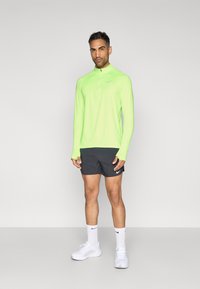 Αθλητικό τοπ με μακριά μανίκια σε νέον πράσινο χρώμα, με γιακά και φερμουάρ μέχρι το ύψος του στήθους, σε συνδυασμό με μαύρες σορτς και λευκά αθλητικά παπούτσια· διαθέτει λογότυπο Nike.