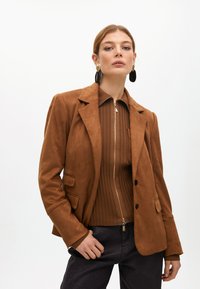 Hnědý semišový blazer s klopami, dvěma předními kapsami a volným střihem, vrstvený přes hnědé žebrované tričko se zipem.