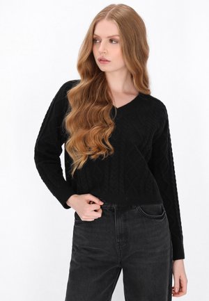 Kvinde med langt bølget blondt hår iført en sort striktrøje og sorte jeans, poserer med den ene hånd ved taljen, mens hun kigger sidelæns.