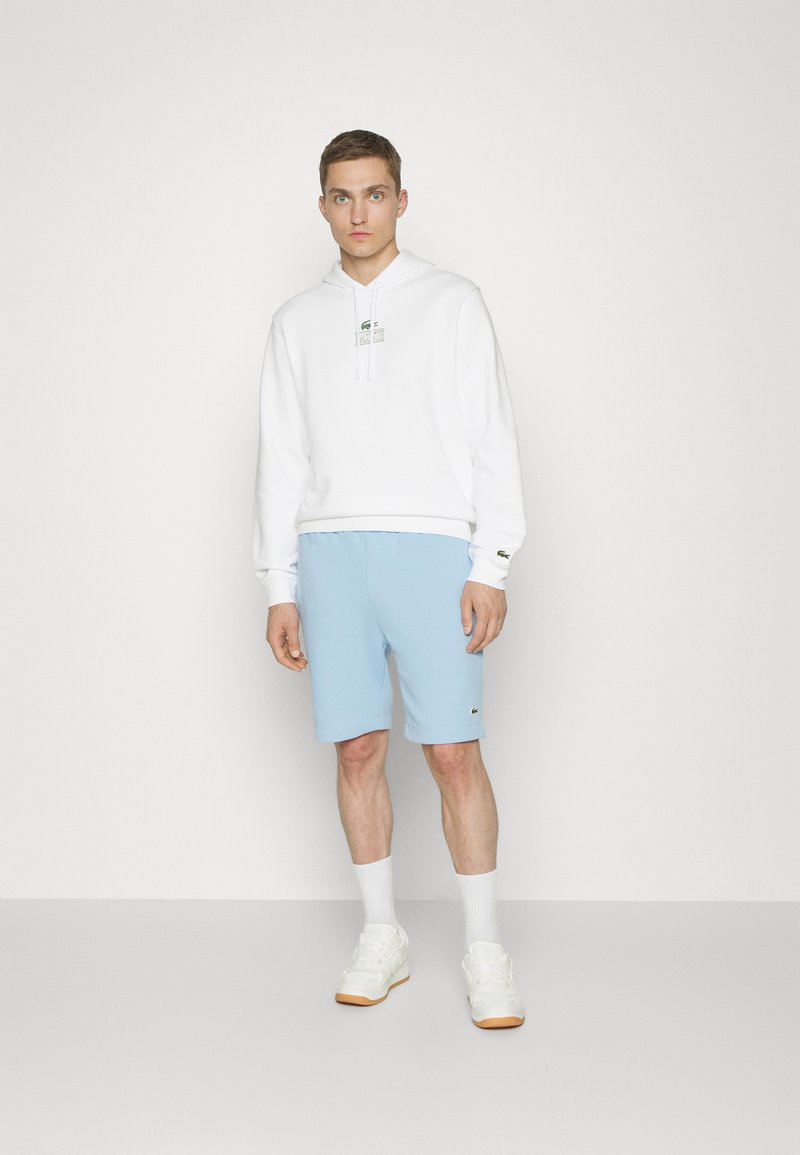 Sweat-shirt à capuche blanc avec un logo vert sur la poitrine, associé à un short bleu clair avec des détails de logo. Porté avec des baskets et des chaussettes blanches.