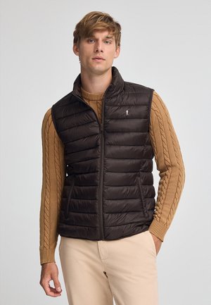 Gilet imbottito marrone con colletto alto, chiusura a zip frontale e trapuntatura orizzontale. Indossato sopra un maglione a treccia, con un piccolo logo ricamato.