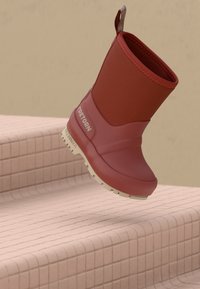 Bordowe wodoodporne buty z beżową podeszwą i logo Tretorn, umieszczone w powietrzu nad różowymi, kafelkowymi schodami.