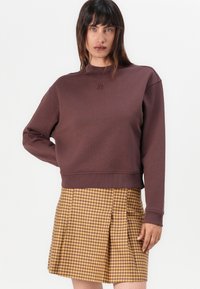 Brun cropped genser med ribbestrikkede mansjetter og krage, med et preget logo. Kombinert med et plissert skjørt i gult og brunt rutemønster.