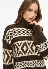 Rjav in krem pulover z visokim ovratnikom in geometrijskimi vzorci z črnimi poudarki. Mehka tekstura in sproščen kroj.