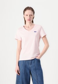 Rosa v-hals t-skjorte laget av myk bomull, med korte ermer og en liten blå logo på venstre bryst. Stilt med vide denim-bukser.
