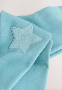 Tessuto azzurro chiaro con una texture morbida, caratterizzato da una stella ricamata in materiale peluche. Il design include una sezione piegata.