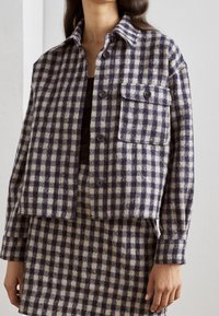 Veste en flanelle à carreaux bleu marine et crème, avec un col, une fermeture à boutons et une poche poitrine. Tissu doux et texturé.