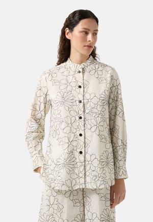 Bloes met bloemenpatroon van lichte stof. Heeft zwartomlijnde bloemen op een crèmekleurige achtergrond, met een mandarin kraag en knoopsluiting.