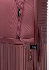 Valise rigide de couleur rose foncé, présentant des rainures verticales texturées, des double zippers, un cadenas à combinaison et un logo de marque bien en vue.