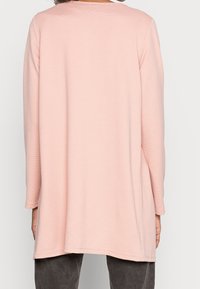 Person som bär en långärmad, ljusrosa ribbad topp med en lös passform, sedd bakifrån, i kombination med mörkgrå byxor.