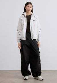 Λευκό τζάκετ με φερμουάρ και μαύρες λεπτομέρειες, με δομημένο γιακά. Φορεμένο πάνω από μαύρη μπλούζα με σχέδια και συνδυασμένο με φαρδιά μαύρα cargo παντελόνια και αθλητικά παπούτσια.