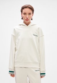 Light beige hoodie with a loose fit, made of smooth fabric. Features green logo and stripes on cuff, with a classic hood design.