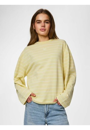 Jeune femme portant une chemise rayée jaune pâle à manches longues et un jean bleu clair, debout les mains dans les poches devant un fond uni.