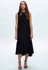 Svart ärmlös klänning med knut i halsen, texturerad tyg, utsvängd nederkant och volangfält. Bärs med svarta klacksandaler.