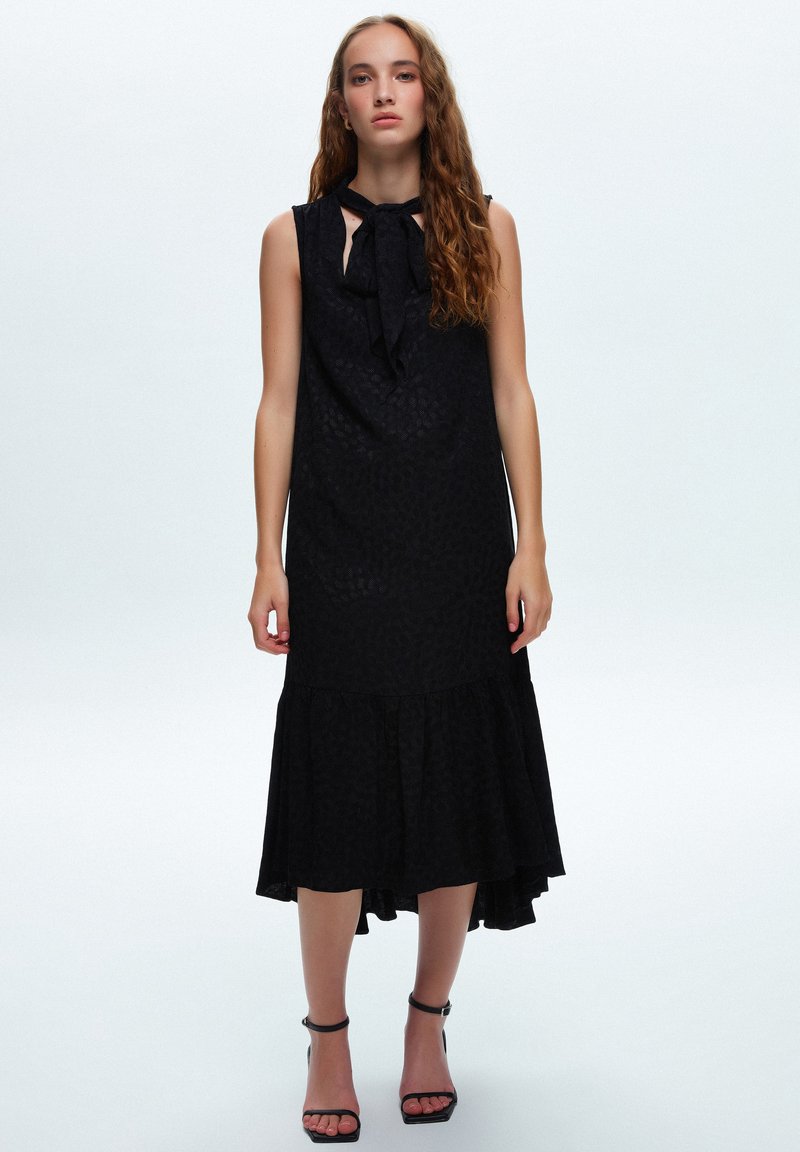 Svart ärmlös klänning med knut i halsen, texturerad tyg, utsvängd nederkant och volangfält. Bärs med svarta klacksandaler.