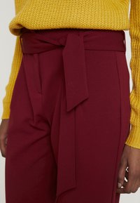 Pantalons larges bordeaux avec une ceinture nouée à la taille. Assortis à un pull en tricot jaune texturé, mettant en valeur des tissus superposés et des couleurs distinctes.