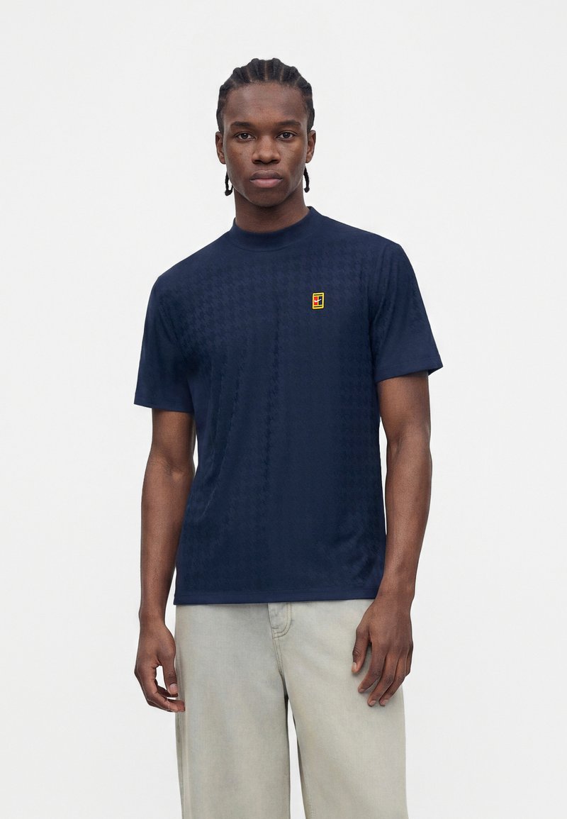 Man with braided hair wearing a navy blue short-sleeve shirt with a small yellow and red logo on left chest, beige pants, standing against white background.