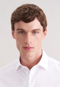 Witte overhemd met knoopsluiting, klassieke kraag, gladde textuur en strakke lijnen, gedragen door een mannelijk model met kort, golvend bruin haar en lichtblauwe ogen.