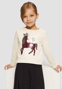 Langärmliges Cremehemd mit einem Pferdemotiv aus mehrfarbigen Pailletten. Kombiniert mit einem schwarzen Faltenrock.