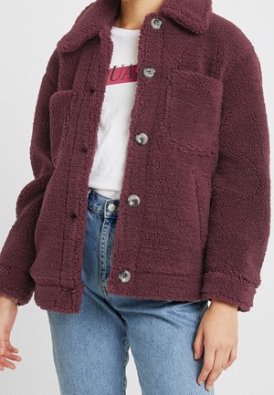 Personne portant une veste polaire texturée violet foncé boutonnée par-dessus un t-shirt blanc avec un motif graphique et un jean bleu clair.