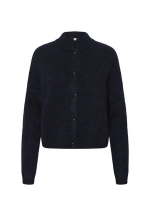 Femme aux cheveux bouclés portant un cardigan noir boutonné, un jean bleu large avec revers et des talons noirs pointus, debout devant un fond blanc.