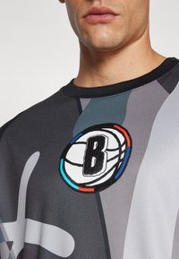 Basketball-tema grå trøje med sort krave, der har et cirkulært logo med et stiliseret "B" og farverige accenter på brystet.