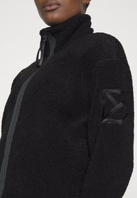 Czarna kurtka polarowa z wysokim kołnierzem, zamkiem błyskawicznym z przodu i teksturowaną powierzchnią. Posiada geometryczną łatkę z tkaniny na górnej części rękawa.