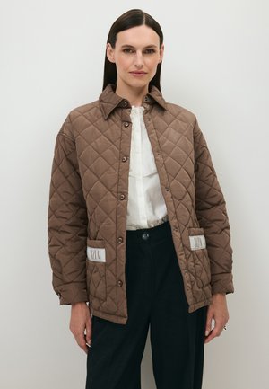 Vrouw met steil donker haar die een bruine gewatteerde jas, een witte blouse met knopen en een zwarte broek draagt, staand tegen een effen achtergrond.