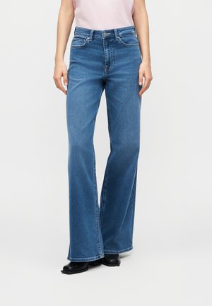 Ženska, oblečena v modre široke kavbojke z visokim pasom, črne gležnjarje in svetlo rožnato obleko znotraj hlač, stoji ob belem ozadju.