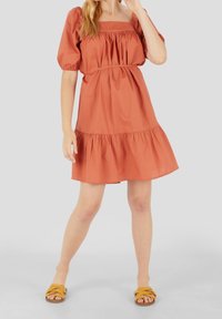 Robe courte orange rouille avec manches bouffantes, encolure carrée et ourlet à volants, associée à des sandales jaune à enfiler. Tissu lisse, coupe décontractée.