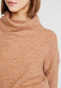 Lichtbruine, gebreide coltrui met een zachte textuur. Heeft een omgevouwen kraag en een geribd patroon, wat warmte en comfort benadrukt.