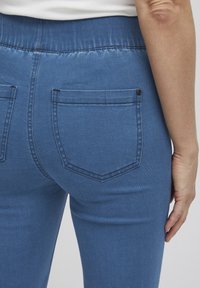 Jeans de mezclilla azul con una forma ajustada, cintura elástica y dos bolsillos traseros con un detalle de un solo botón. Textura suave, diseño de tiro medio.