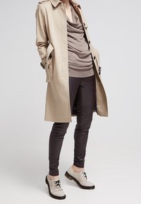 Beige trenchcoat met schouderstukken over een bruine coltrui, gecombineerd met donkere legging en lichtgrijze veterschoenen.