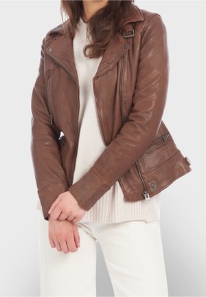 Frau trägt eine braune Lederjacke mit Reißverschluss-Details, einen weißen Strickpullover und weiße Hose, steht mit sanft verschränkten Händen.