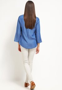 Jasnoniebieska koszula denimowa z rękawami 3/4 w kształcie dzwonu, zestawiona z białymi obcisłymi dżinsami i brązowymi butami, widok z tyłu.