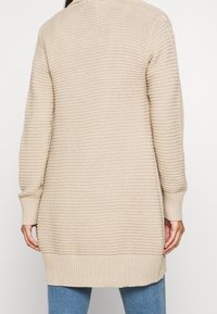 Beige lange, gerippte Strickjacke mit umgeschlagenen Ärmeln, getragen über blauen Jeans, von hinten gezeigt.