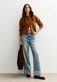Kvinna i brun kofta, vit crop top, vida jeans med uppvikt fåll och bruna lägenheter, med en brun väska mot en enfärgad vägg.