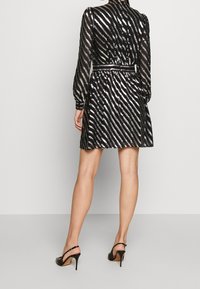 Robe noire avec des rayures diagonales argentées, manches longues, taille ceinturée et jupe fluide. Sandales à talons hauts noires. Tissu lisse et texturé.