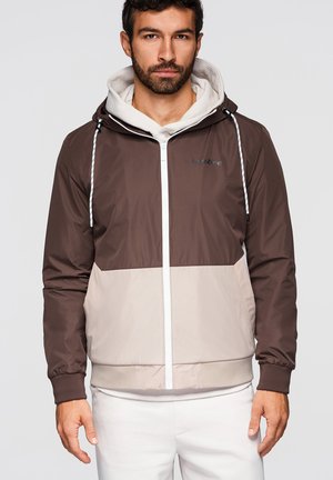 Mand iført brun og beige hættejakke med lynlås over en lys hættetrøje, stående mod en ensfarvet lys baggrund, vendt mod fronten.