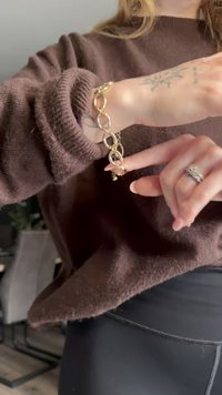 Zelta ķēdes aproce ar savienotām ovālām saitēm un nelielu piekariņu, uzvilkta virs brūnas džemperis. Tekstūra izskatās gluda un pulēta.