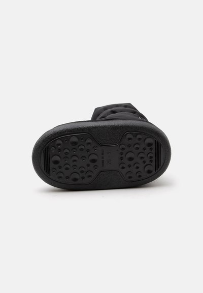 Semelle de botte noire avec des grips circulaires texturés et en relief ainsi que des indications de taille, présentée sur un fond clair uni.